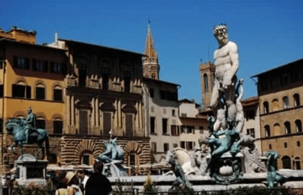 Statues, including a large Neptune figure, stand in an historic Italian square surrounded by old buildings and a church tower under a clear blue sky.