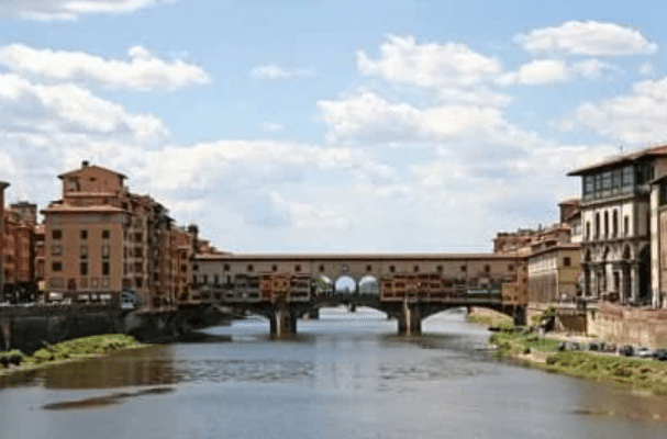 A historic stone bridge with buildings on top spans a river between two rows of old city buildings under a partly cloudy sky.