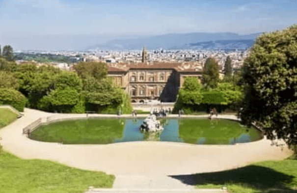 A formal garden with a pond and statue in front of a historic building; cityscape and mountains visible in the background under a clear sky.