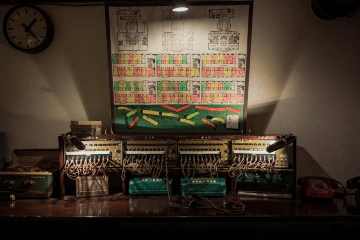 A dimly lit desk with vintage switchboards, tangled wires, a wall clock, a red rotary telephone, and a board with diagrams and colour-coded charts above.