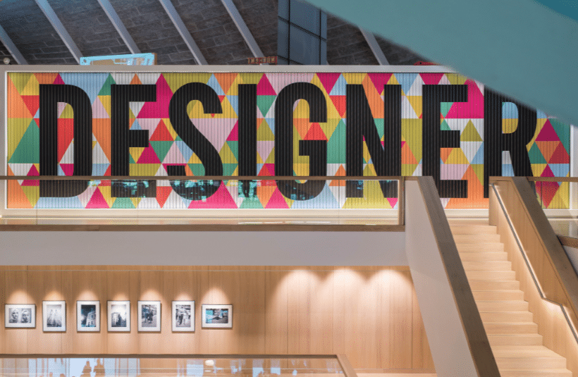 A colourful wall with the word “DESIGNER” in large black letters and geometric patterns above a wooden staircase and framed photos.
