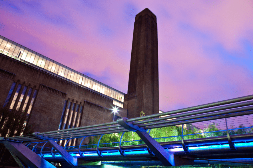 The image shows the Tate Modern art museum’s brick tower and adjacent Millennium Bridge under a purple and blue evening sky in London.
