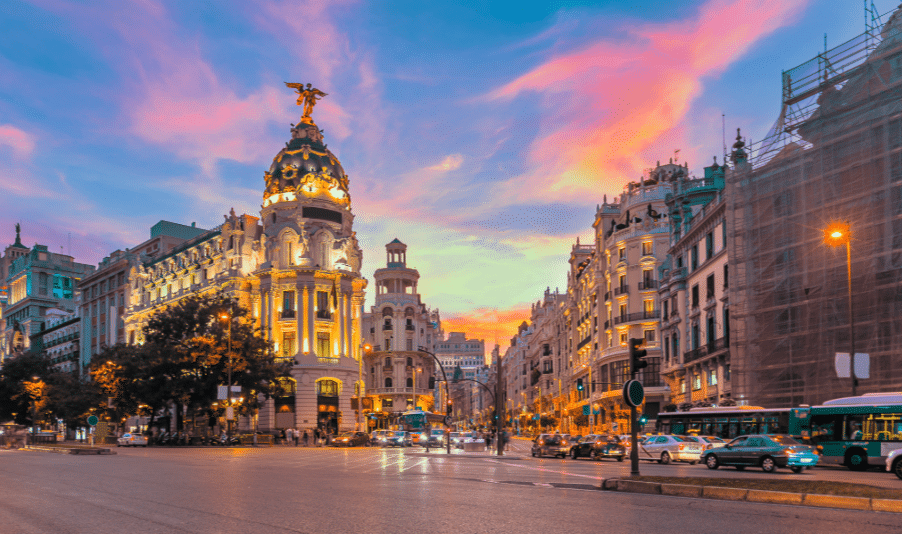 City street in Madrid at sunset with the Metropolis Building lit up, surrounded by cars, buses, and pedestrians under a colourful sky.