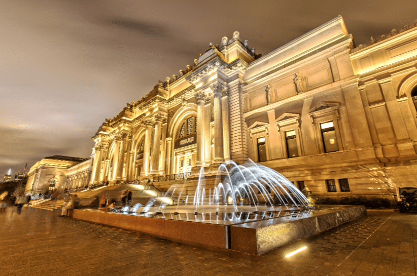 A large, illuminated classical-style building at night with a central fountain in front and blurred figures walking nearby.