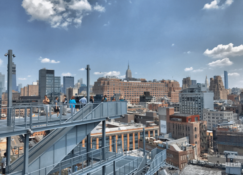 People stand on an outdoor observation deck overlooking a city skyline with tall buildings under a partly cloudy sky.