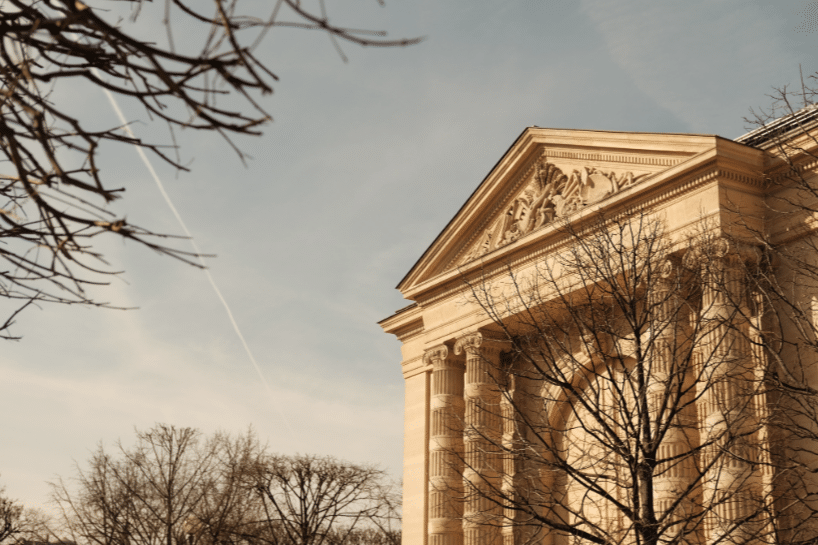 A neoclassical building with tall columns and ornate pediment, partially framed by leafless tree branches under a clear sky.