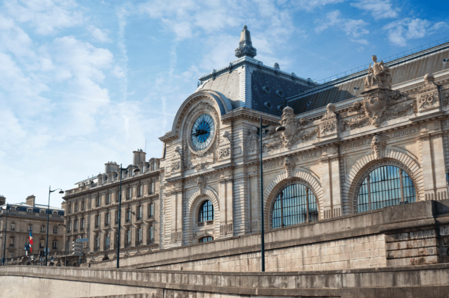 Stone building with arched windows and a large clock on the facade, set against a blue sky with scattered clouds.