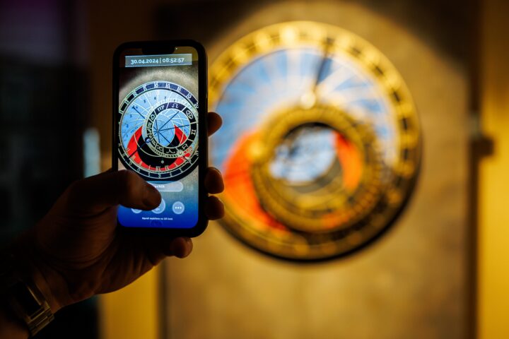 A hand holds a mobile phone displaying a digital image of an astronomical clock, with the actual clock blurred in the background.