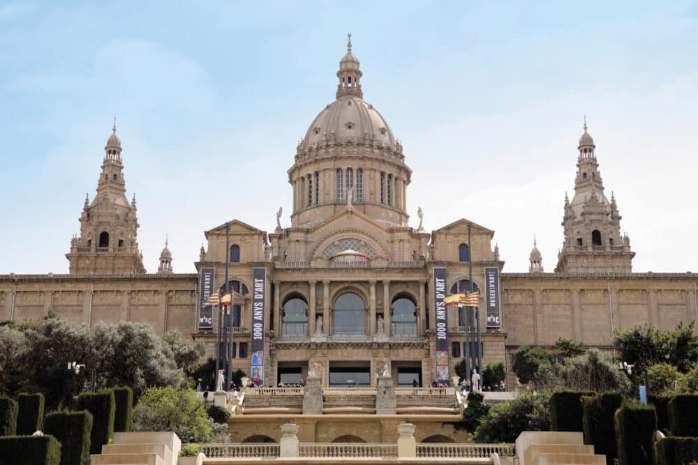 The image shows the front view of the Museu Nacional d’Art de Catalunya in Barcelona, with steps, greenery, and clear sky in the background.