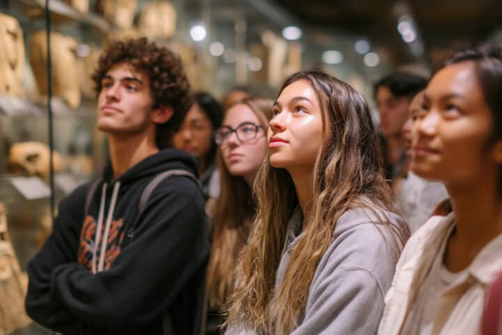 A group of young people stand closely together in a museum, attentively observing artefacts displayed in glass cases.