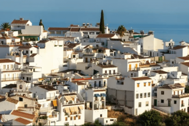 Cluster of white buildings with red-tiled roofs on a hillside overlooking the sea under a clear sky.
