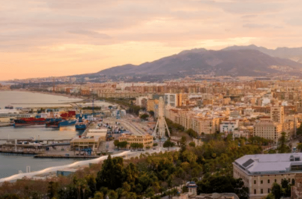 A coastal cityscape shows a harbour with ships, a Ferris wheel, and urban buildings, with mountains in the background under a cloudy sky at sunset.