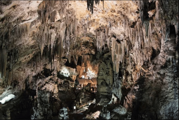Large cave interior with pointed stalactites hanging from the ceiling and stalagmites rising from the floor; people are visible near illuminated structures in the centre.