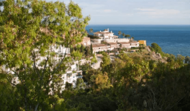 Buildings on a hillside overlook the sea, surrounded by green trees under a partly cloudy sky.